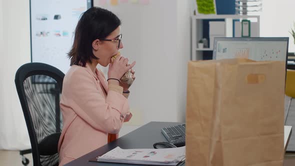 Businesswoman Eating Bite of Tasty Sandwich Drinking Coffee in Front of Monitor