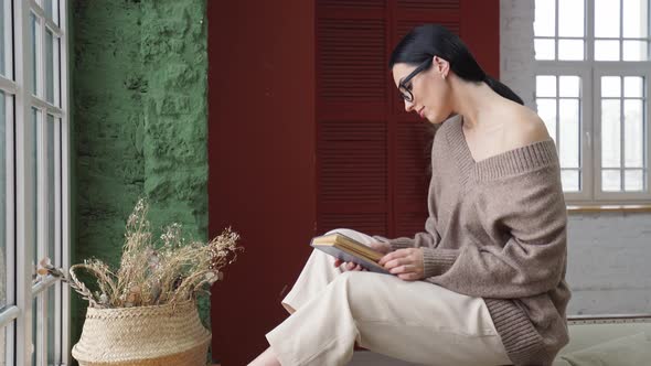 a Young Woman in Cozy Clothes and a Smooth Haircut Reads a Book Sitting By the Window