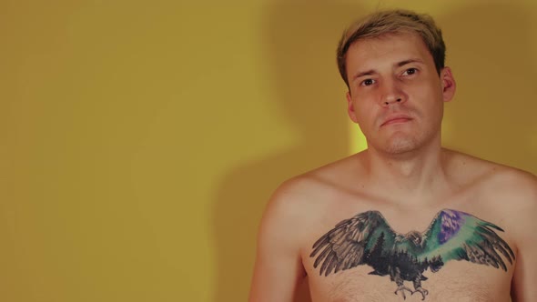 A Handsome Man with a Bare Torso with a Tattoo on His Chest Looks at the Camera Standing on a Yellow