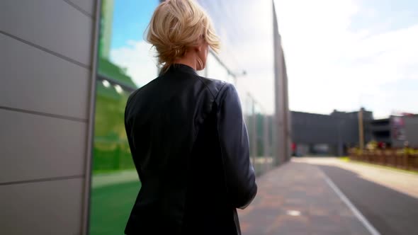 the Blonde Goes Along the Wall with the Big Windows, Turns Around and Takes Off Her Black Jacket