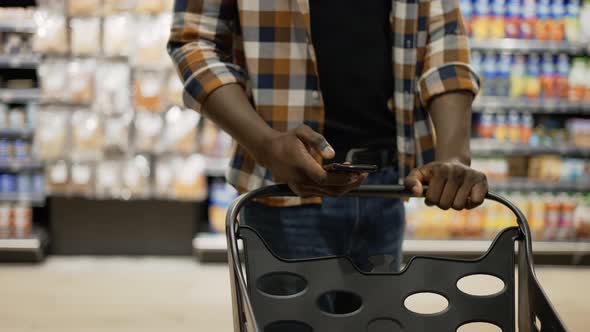 Unrecognizable Man with Smartphone Pushes Shopping Cart Walks at the Store