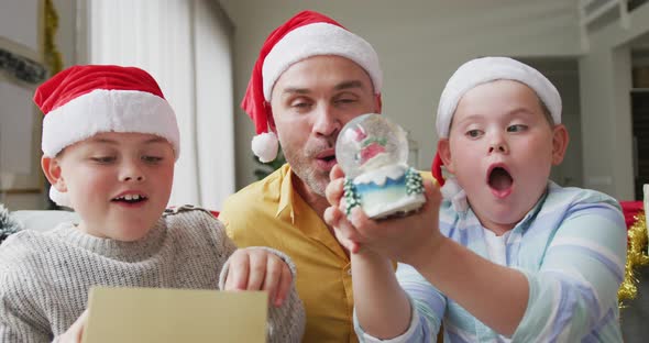 Caucasian father and two sons wearing santa hats holding a snow globe sitting on the couch at home