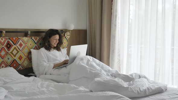 Young Woman Holds Laptop and Types Smiling Sitting on Bed