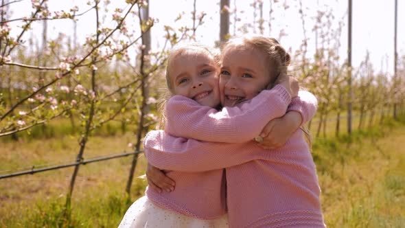 Two Little Twin Sisters Dressed in Identical Dresses Hug in an Apple Orchard