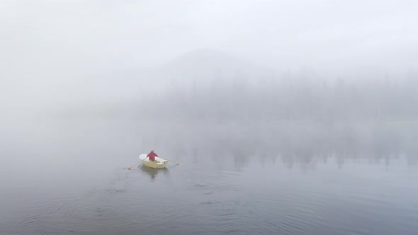 Flying behind a person rowing a small boat on a mist filled lake with forest and mountain in the bac