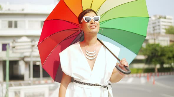 Asian lgbtq guy wearing woman clothes with sunglasses holding rainbow umbrella walking down street.