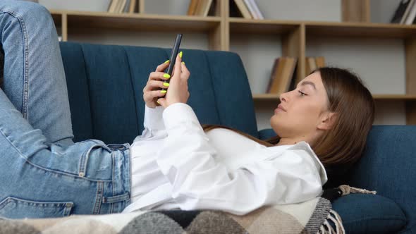 Happy Young Woman Using Mobile Phone While Lying a Couch at Home with Laptop Computer