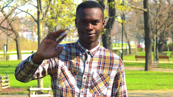A Young Black Man Smiles and Waves at the Camera in a Park on a Sunny Day