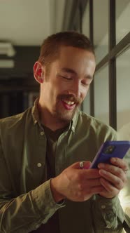 A Young Guystudent Stands Near the Window Looks Far and Then Reads Posts on the Internet Smiling