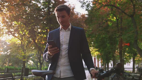 Handsome Young Stylish Man Using Smartphone Outdoors with Bicycle in Park in City Center During