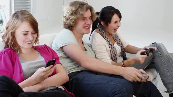 Teenage girl texting on smartphone, friends playing video game