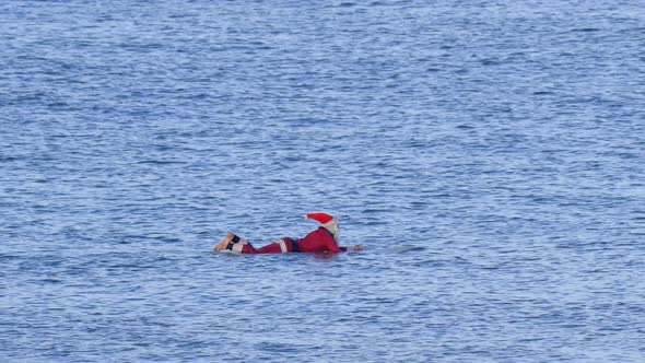 Santa Claus sits on his surfboarding waiting for waves while surfing.