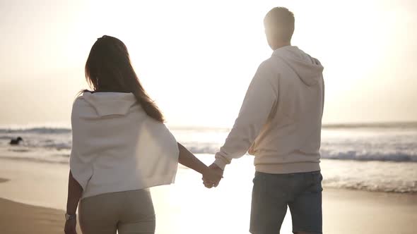 Backside View of Young Couple in Love Holding Hands Walking on the Beach in Casualclothes