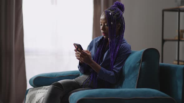 Afroamerican Lady with Modern Smartphone is Resting at Home Sitting Alone on Couch and Reading News