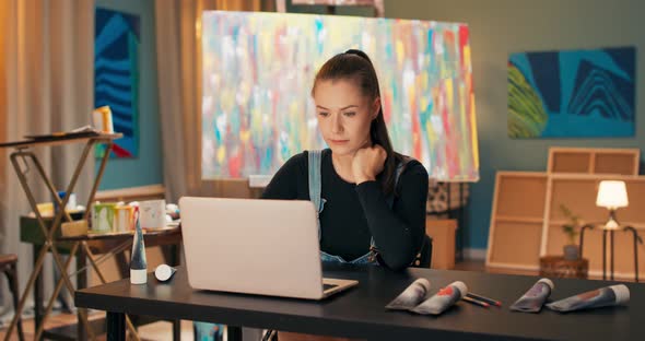 A Smiling Brunette Sits at a Desk in Front of a Laptop in Art Studio