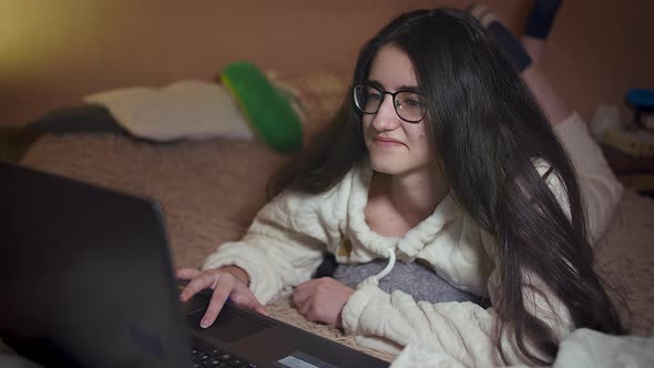 Smiling girl with glasses lying on the bed at night near the laptop, communicates in social networks