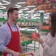 Friendly handsome male stock clerk in red apron, helping young attractive woman to choose fruits  - VideoHive Item for Sale