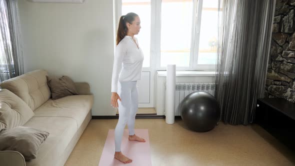 Girl in White Tracksuit Does Squats on Mat in Light Room