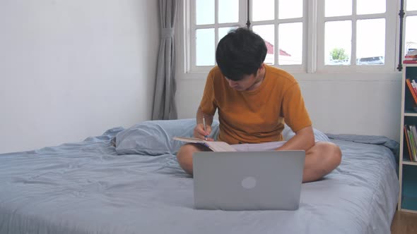 Man Working With A Laptop And Writing In Book On The Bed