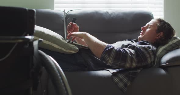 Happy caucasian disabled man lying on sofa in living room using laptop