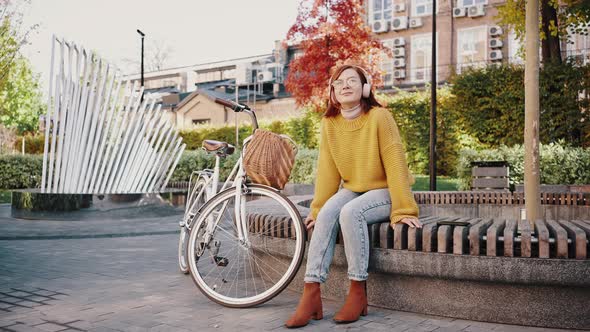 Young Female Sitting on Bench in City Park Listening to Music Through Headphones and Smiling