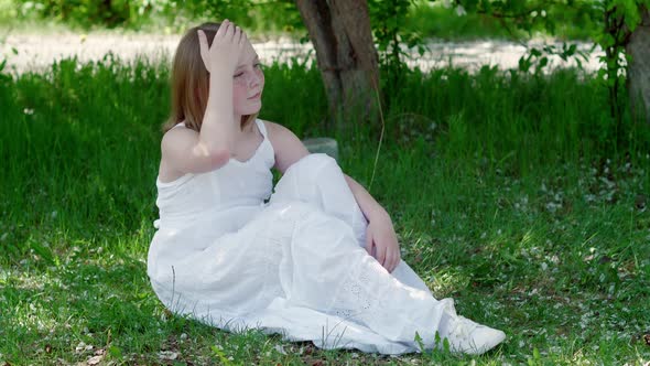 Relaxed Girl in White Dress Sitting on Green Grass in Summer Garden