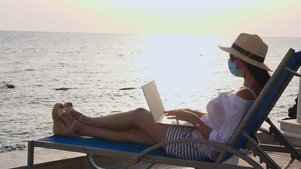 Young Woman in Protective Mask, Sun Hat and Summer Clothes, Working on Laptop, Sitting on Sun