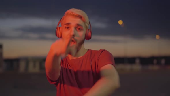 Carefree Caucasian Man in Headphones Dancing in the Evening Outdoors in Multicolored Laser Lights