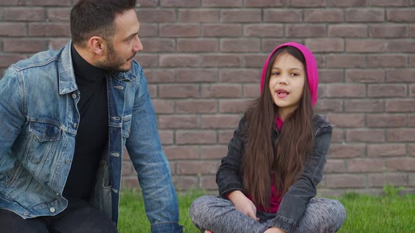 Dad and Daughter Are Sitting, Having Fun and Chewing Bubble Gum Against the Backdrop of Grass and a