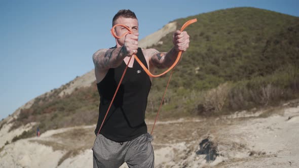 Muscular Tattooed Sportsman in Black Tank Top Trains with Elastic Bands Outdoor