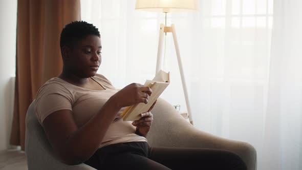 PlusSized African Lady Reading Book Sitting In Chair At Home