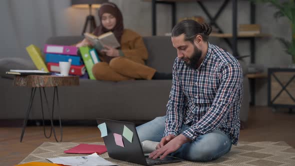 Wide Shot of Middle Eastern Man Sitting on Floor in Living Room with Laptop and Blurred Woman in