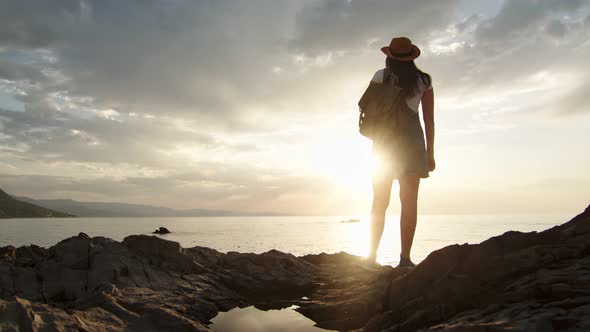 Back View Travel Female with Backpack Standing on Top of Mountain Over Sea Surrounded By Sunlight