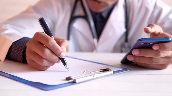 Doctor Hand Writing Prescription and Using Smart Phone on Clinic Desk