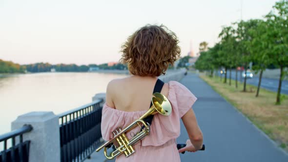 the Happy Female Musician Rides a Scooter Along the Promenade with a Trumpet Behind Her Back