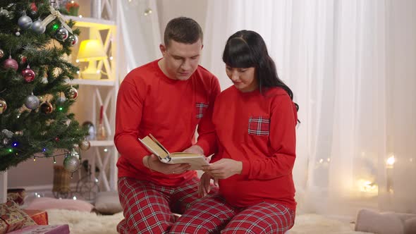 Portrait of Cheerful Couple in Similar Red Pajamas Reading Book Together Sitting at Christmas Tree