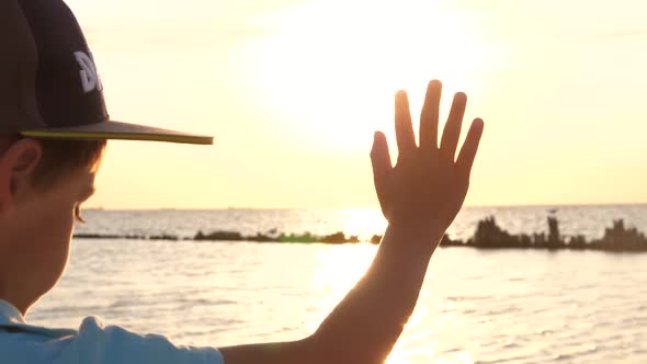 A Little Boy Stretches Out His Hand To Catch the Rays of the Sun As It Sets Over the Sea. Close Up.