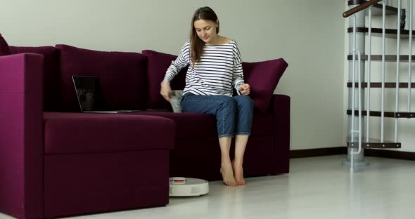 Closeup of robot vacuum cleaner driving around barefoot woman working at home office. 