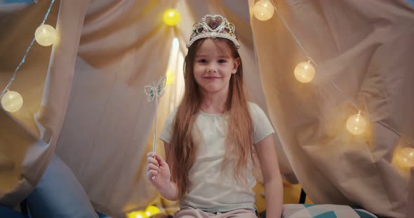 Portrait of Adorable Little Girl Wearing Crown and Holding Magic Wand Sitting in Teepee Tent at Home