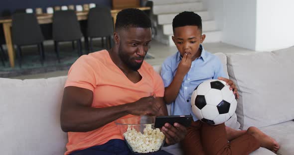 African american father and son eating popcorn and watching a football game on smartphone