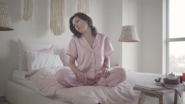 Portrait of Relaxed Caucasian Woman Sitting on Bed in the Morning. Brunette Lady in Pajamas Waking