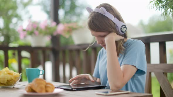 Teenage Girl Sits At Wooden Table In a Summer Cafe In Headphones With Phone