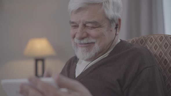 Close-up of Laughing Senior Caucasian Man in Earphones Watching Series of Film on Smartphone