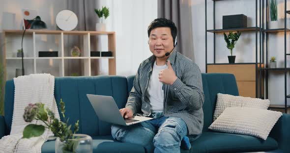 Asian Male Worker in Headphones Sitting on the Couch at Home Office and Looking into Camera