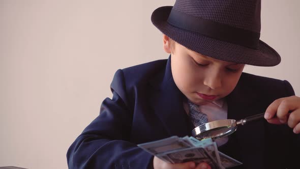 Child Boy Looks Like a Businessman in Hat and Suit is Looking at Dollars with Magnifier