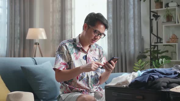 Asian Young Man Using Smart Phone While Packing Clothes In Suitcase At Home, Preparing For Vacation