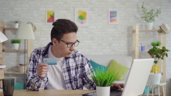 Frustrated Young Asian Man in a Shirt Enters Data with a Bank Card on a Laptop