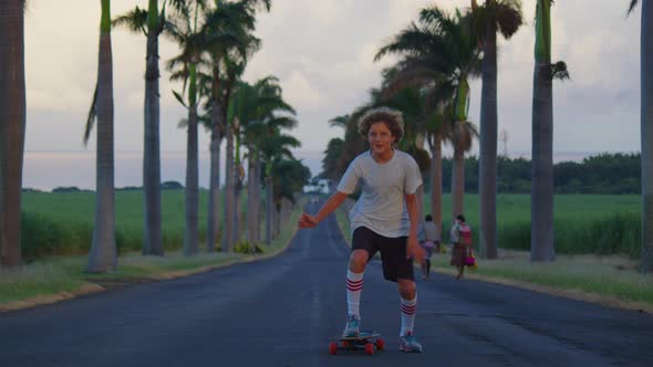 A Teenager with Long Hair Rides a Skateboard Along a Beautiful Road with Palm Trees