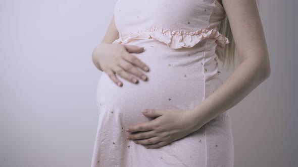 Pregnant Woman in Pink Frock Stands Near White Wall Closeup