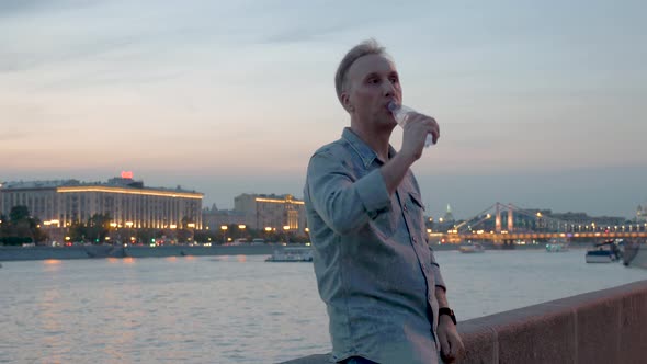 Man Checks Time Drinking Water Evening River and Cloudy Sky